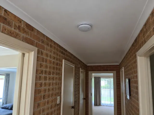 Ceiling smoke alarm in a brick-lined hallway with white ceiling and doorways in Toukley